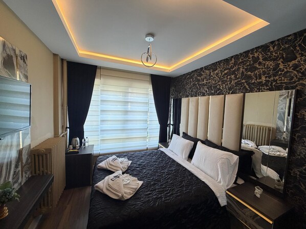 A modern hotel room with a large bed, a bedside table, a mirror, and a window with blinds. - SUITES NECATİBEY (Ankara)