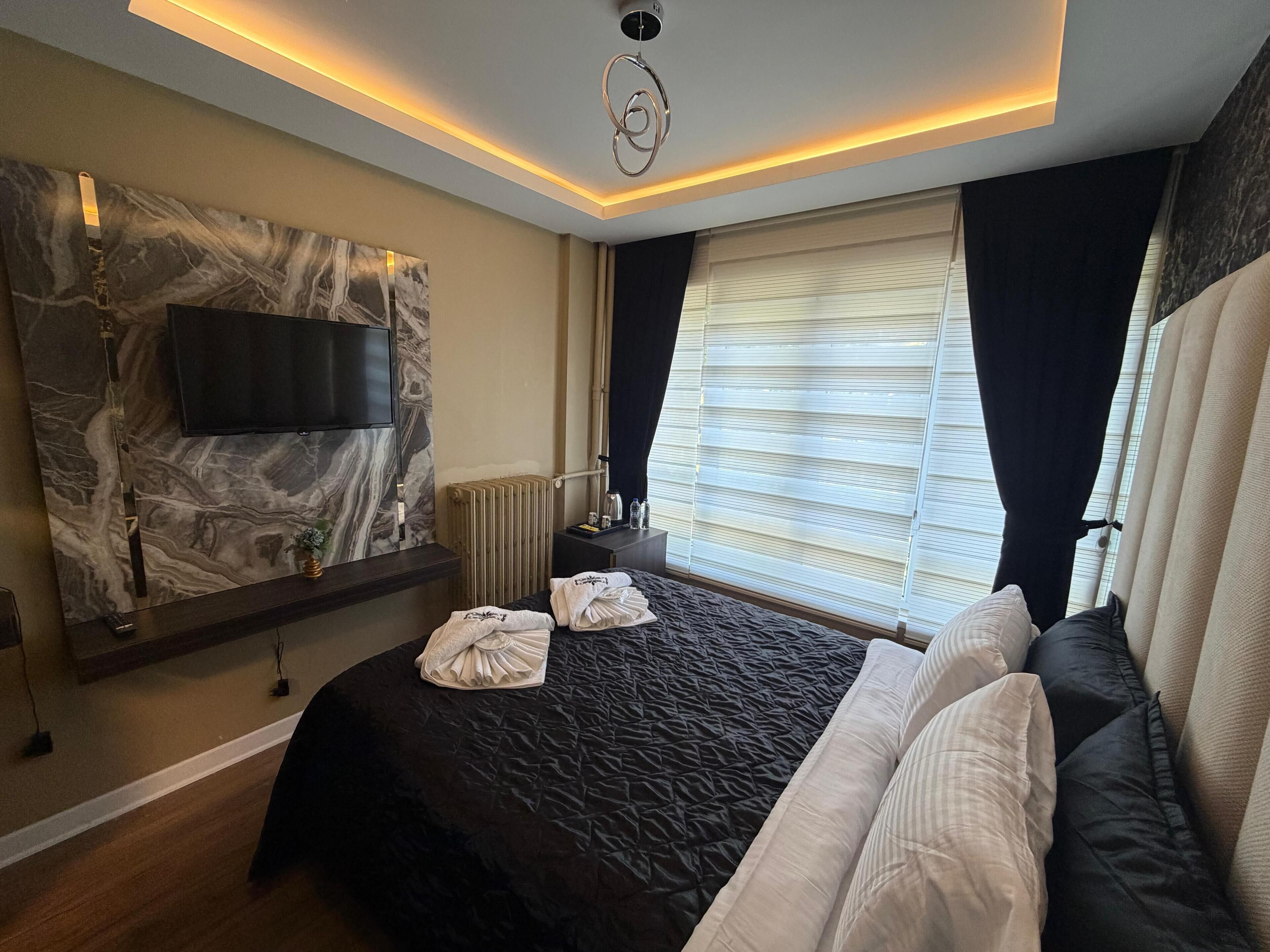 Une chambre d’hôtel moderne dotée d’un grand lit, d’un téléviseur à écran plat fixé sur un mur en marbre, d’un radiateur et d’une fenêtre avec des stores.
