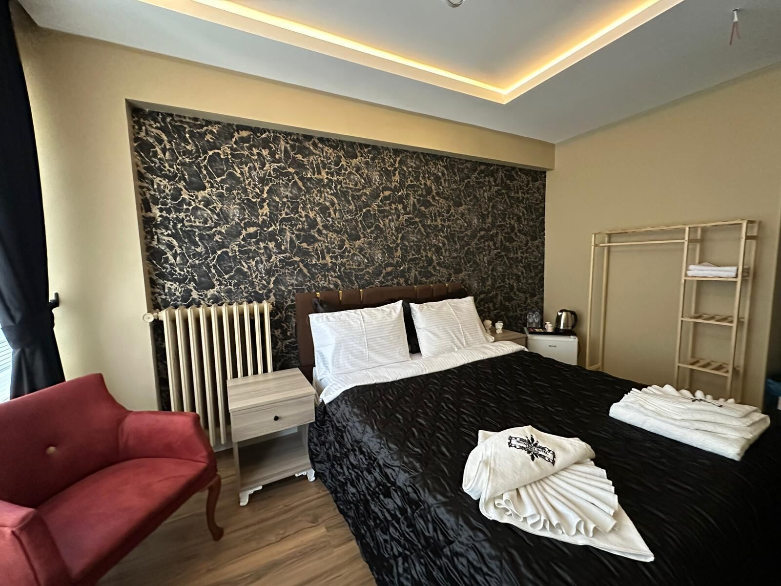 A modern hotel room with a large bed, a red chair, a radiator, and a wardrobe.