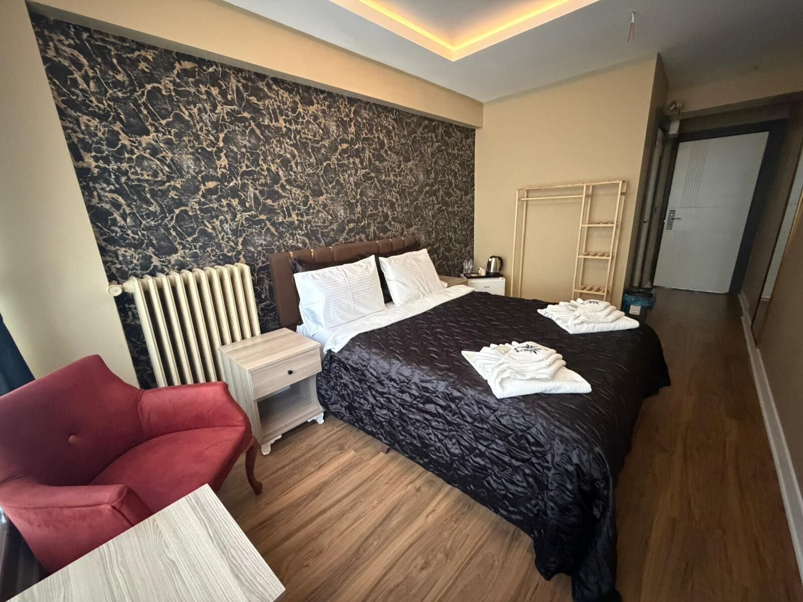 Moderni hotellihuone, jossa on suuri sänky, punainen nojatuoli, yöpöytä ja marmorikuvioitu seinä.