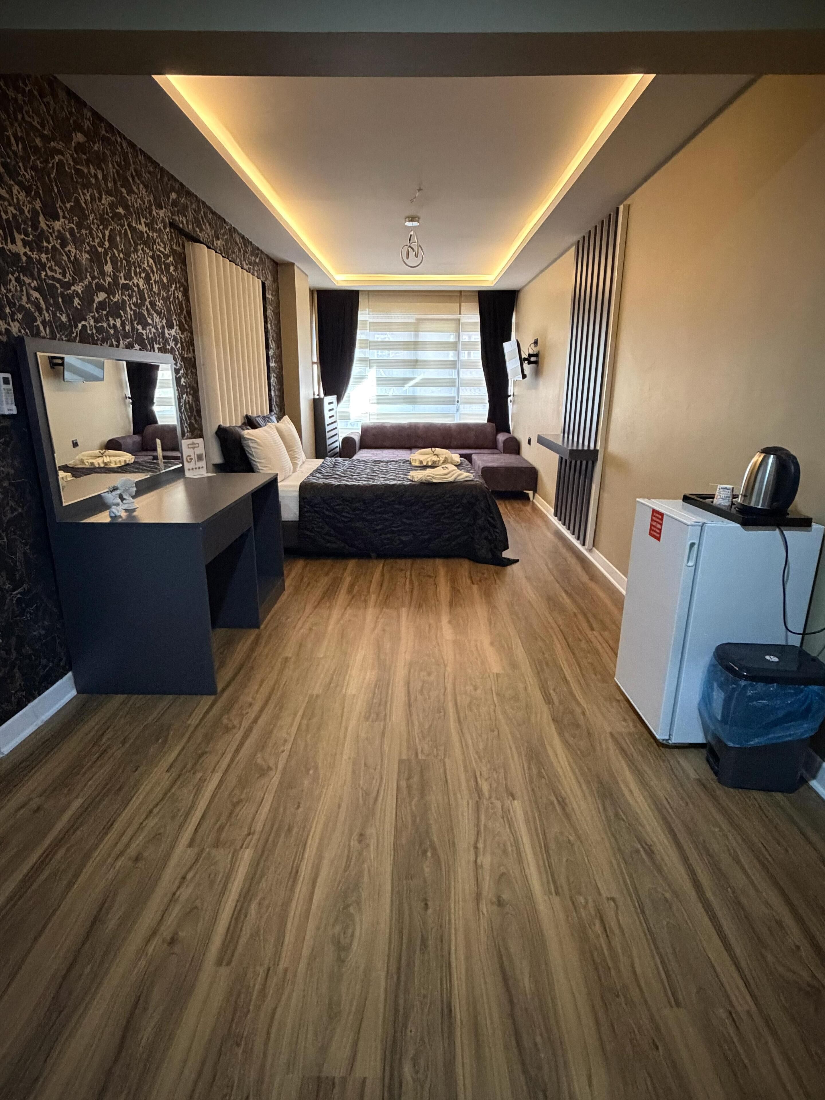 A modern hotel room with a wooden floor, a bed with a dark cover, a desk with a mirror, a small refrigerator, and a TV mounted on the wall.