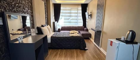 Habitación de hotel moderna con piso de madera, una cama con cubrecama oscura, un escritorio con espejo, un refrigerador pequeño y un televisor montado en la pared.