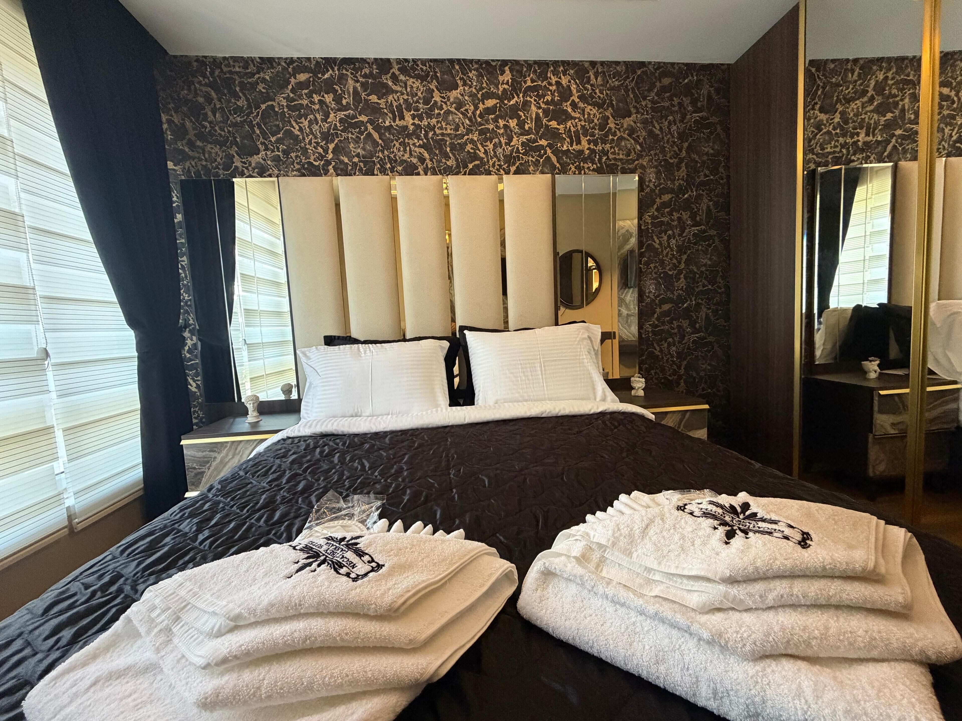 A hotel room with a large bed, a headboard with vertical panels, a bedside table with a lamp, and a mirror on the wall.
