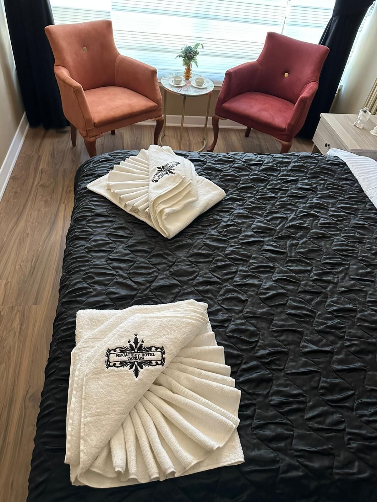 A hotel room with a black quilted bedspread, two red armchairs, a small round table with a plant, and a white towel with a logo on the bed.