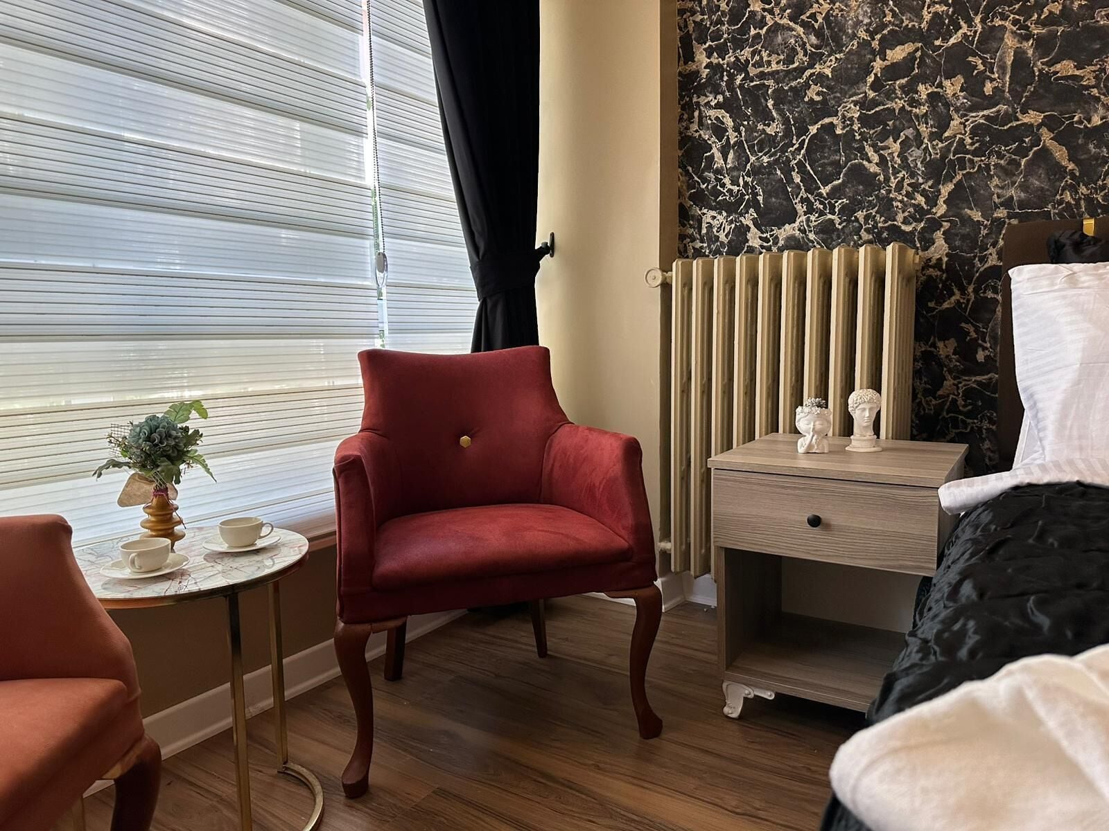 A hotel room with a red armchair, a bedside table with a bust sculpture, a radiator, and a bed with a black and white duvet.