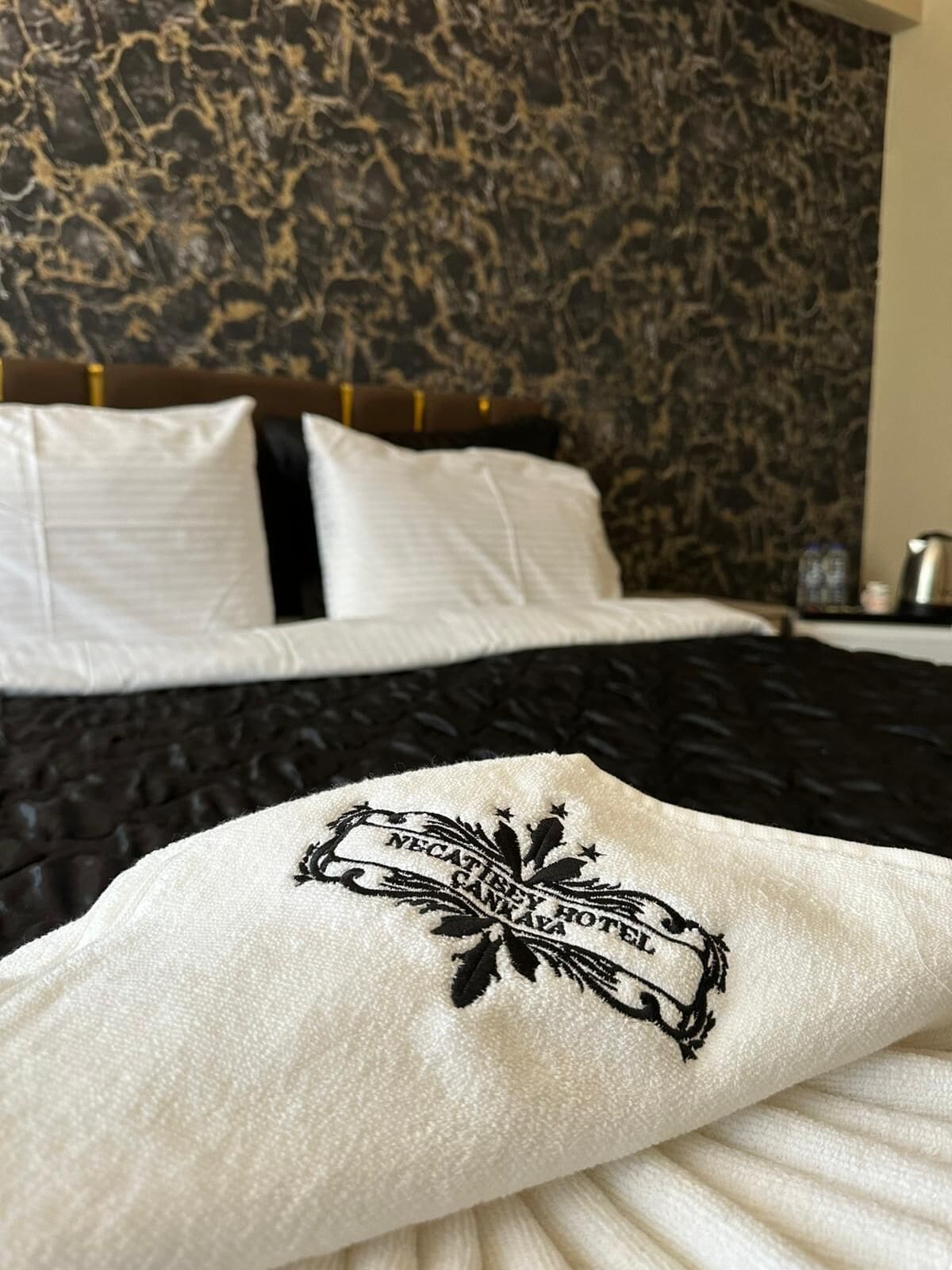 A hotel room with a bed featuring a black and white patterned headboard, a white pillow with a logo, and a textured wall behind the bed.