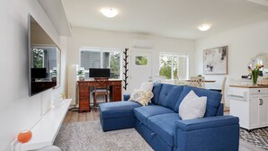 A modern living room with a blue sofa, a desk with a computer, a flat-screen TV, and a dining area. - NEW 2BR Victoria Suite: Family, Pet, Work-Friendly (Colwood)