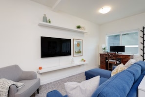 Ein Wohnzimmer mit Flachbildfernseher, einer Couch, einem Schreibtisch mit Computer und einem Fenster.