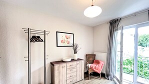 A modern room with a wooden floor, a coat rack, a cabinet with drawers, a chair with a pink blanket, a framed picture on the wall, and a large window with a view of greenery.