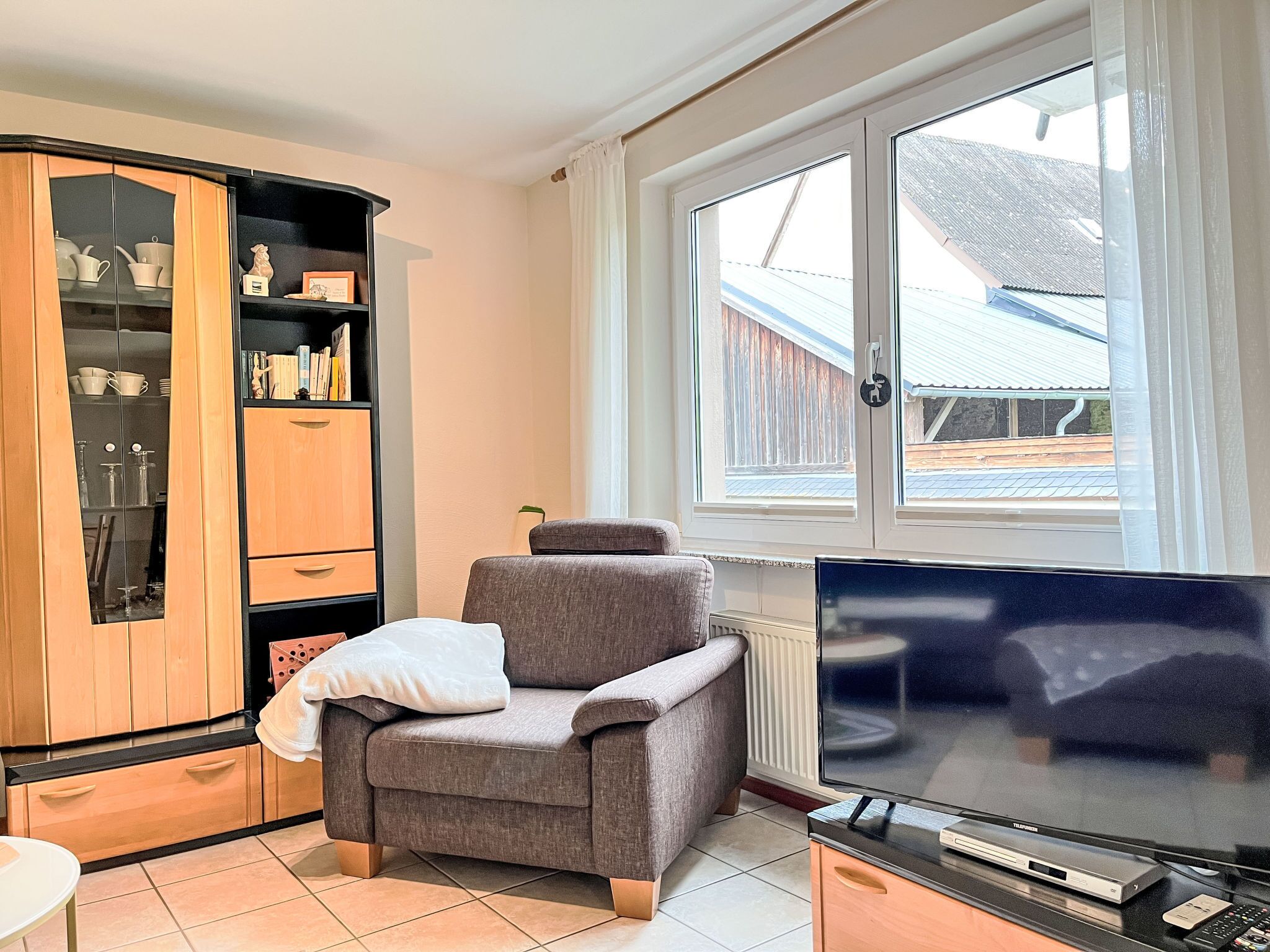 Ein gemütliches Wohnzimmer mit einem grauen Sessel, einer Holzkommode mit Glastüren, einem Flachbildfernseher und einem Fenster mit Blick auf ein Gebäude.