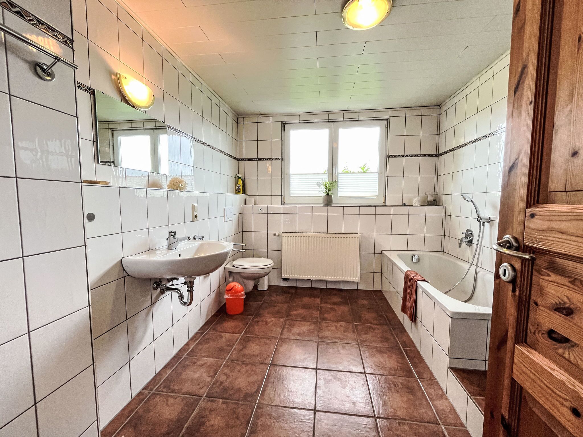 A bathroom with white tiled walls, a brown tiled floor, a sink, a toilet, a bathtub, and a window with blinds.