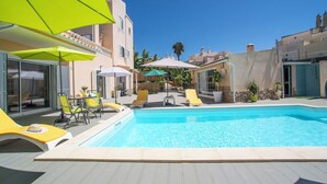 Una piscina con sillones de playa y sombrillas, rodeada de edificios con balcones.