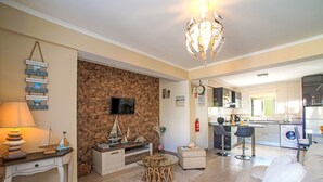Una sala de estar moderna con una pared de acento de piedra, un televisor, un comedor con mesa de vidrio y una cocina al fondo.