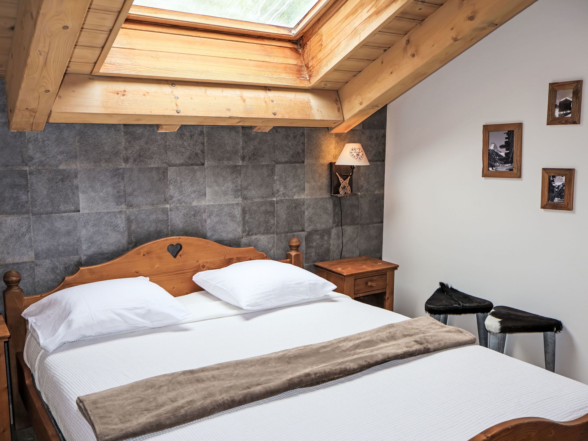 Quarto com teto inclinado, cama com roupa de cama branca, cabeceira de madeira, mesa de cabeceira, abajur e quadros na parede.