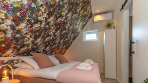 A bedroom with a sloped ceiling, a bed with a pink blanket, white pillows, and a bedside lamp. There is a small window and a plant on the dresser.