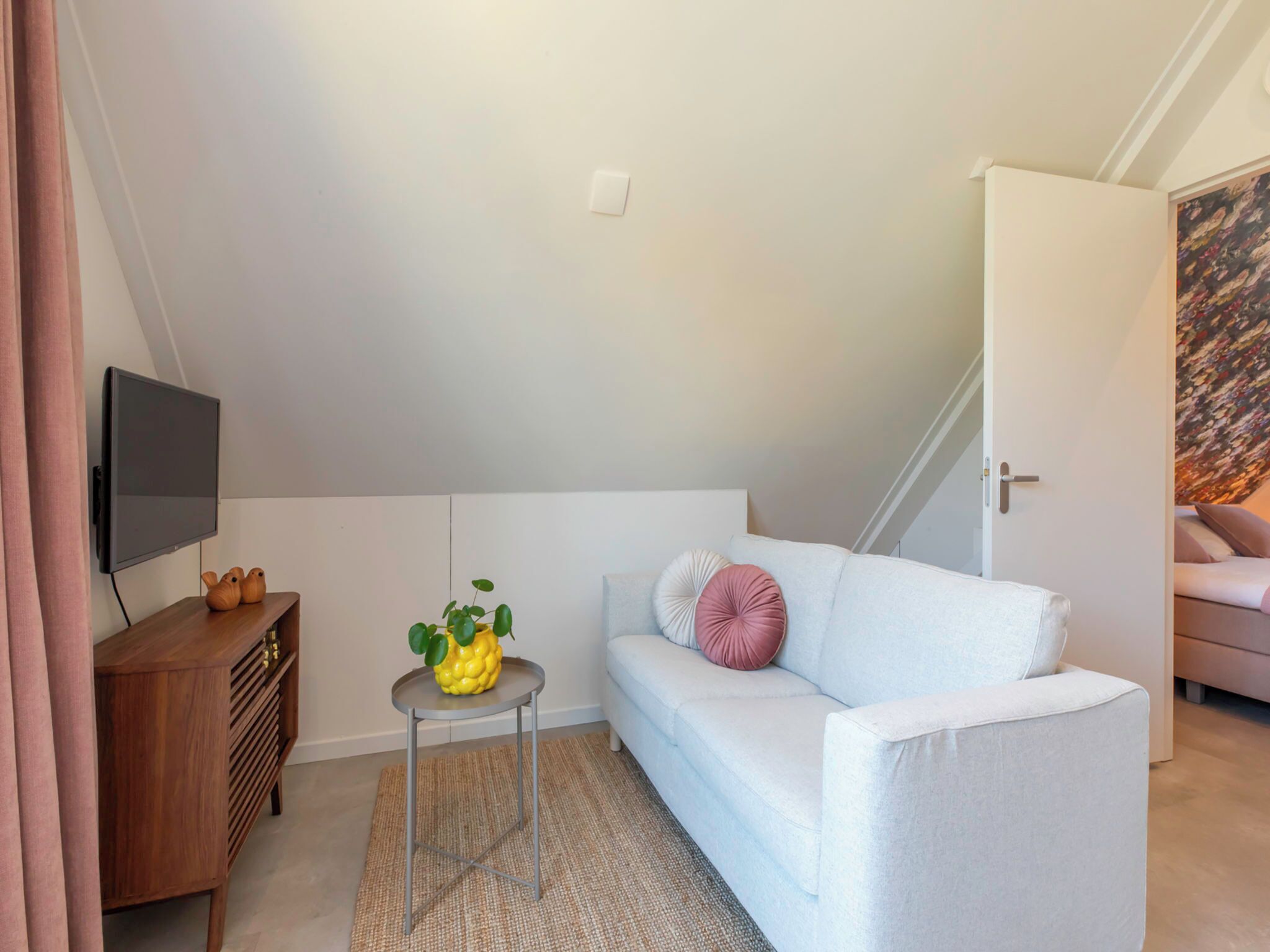 Een moderne woonkamer met een witte bank, een klein bijzettafeltje met een plant, een houten kast en een wandgemonteerde televisie.