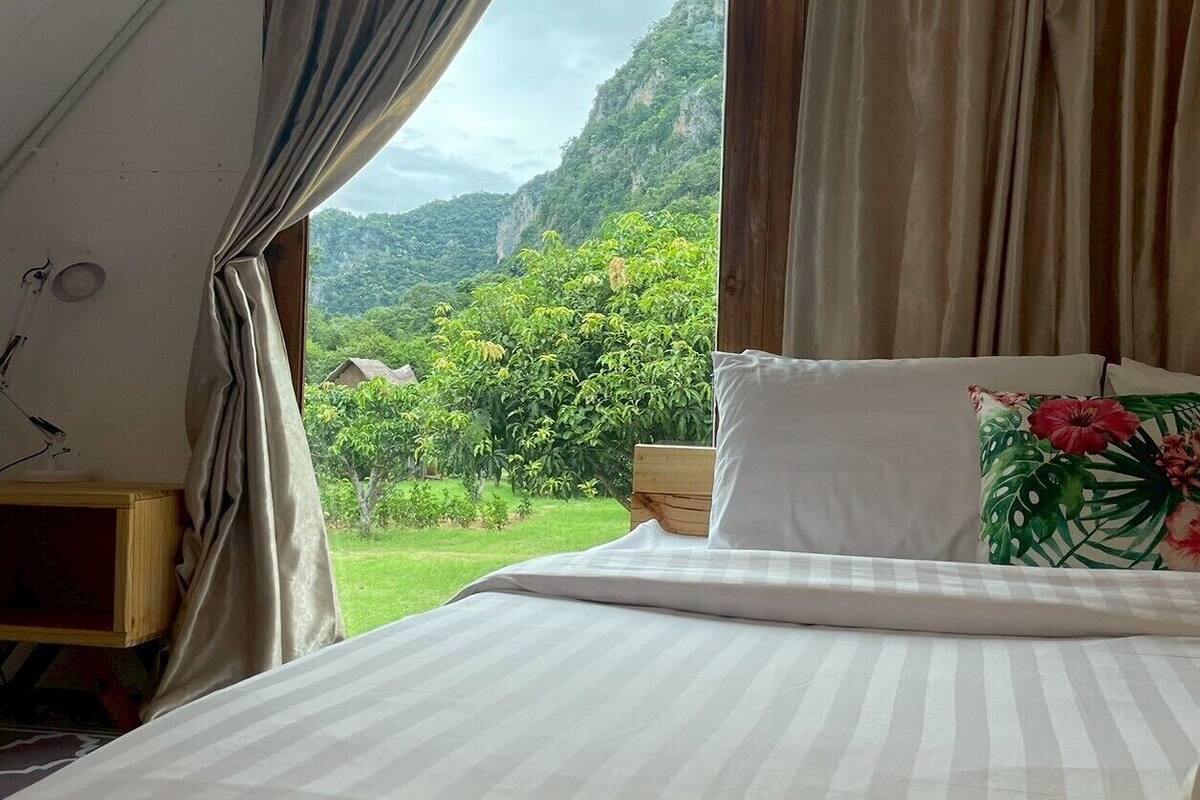 A bedroom with a bed, curtains, a nightstand, and a view of a mountainous landscape.