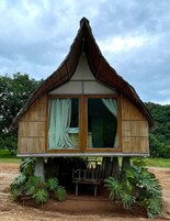 Una pequeña casa de madera con techo de paja, ventanas grandes con cortinas y una mesa bajo el porche.