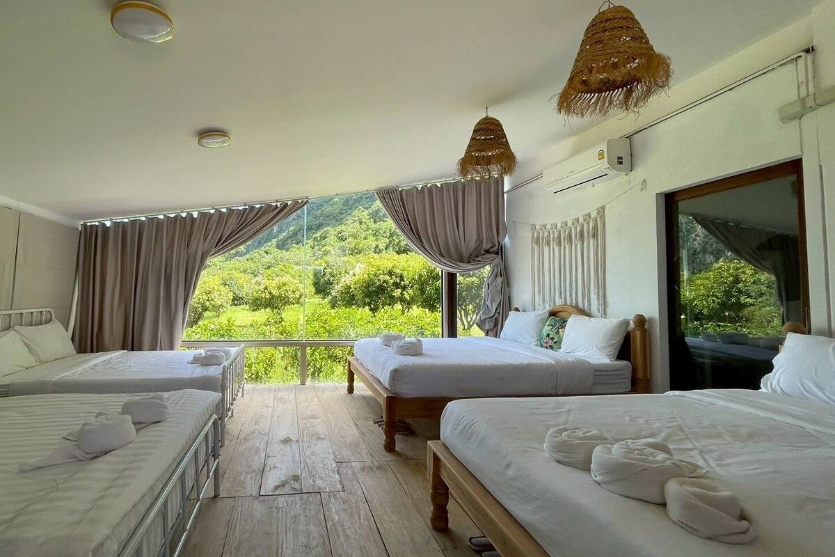 A hotel room with two beds, a large window with curtains, a wooden floor, and a view of greenery.