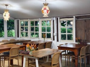 Un vaste espace dédié à la salle à manger, avec des tables et des chaises en bois, de grandes fenêtres et des luminaires suspendus.