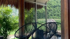 A covered patio area with two black wireframe chairs, a wooden floor, and a view of greenery through large windows. - NUBDAO 168 Homestay (Lan Sak)