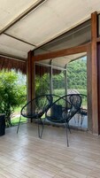 Un patio techado con dos sillas de estructura metálica negra, piso de madera y vista a follaje a través de amplios ventanales.