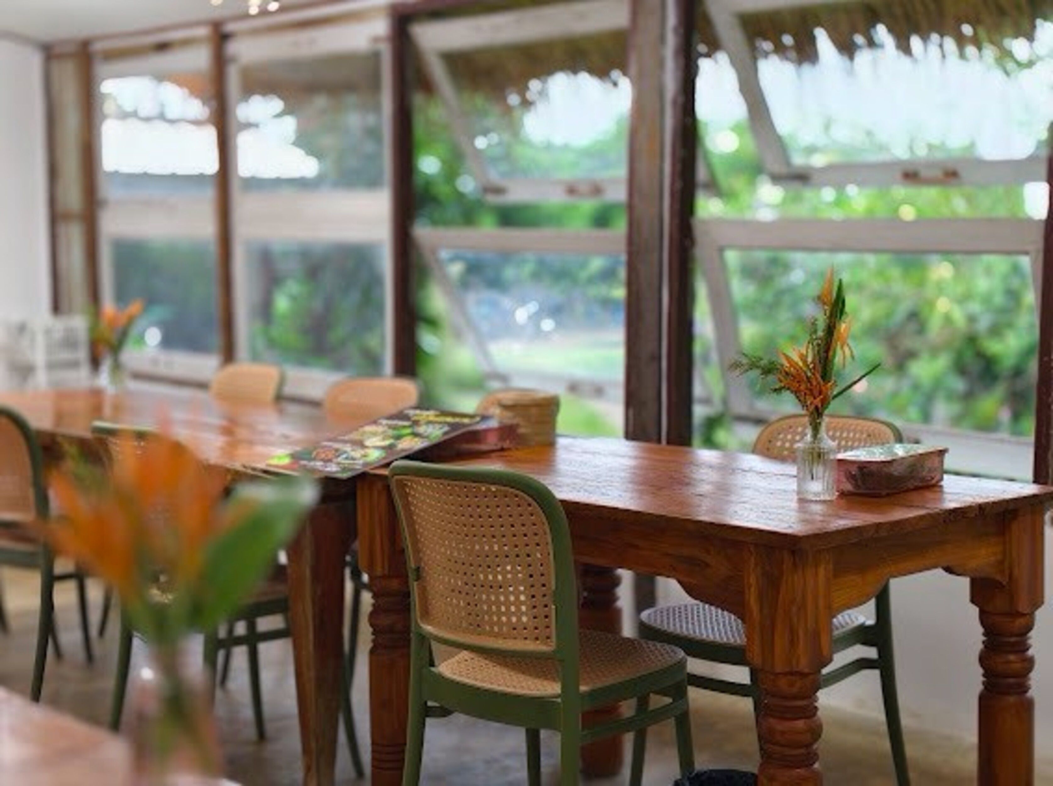 A dining area with a wooden table, wicker chairs, and a view of greenery through large windows.