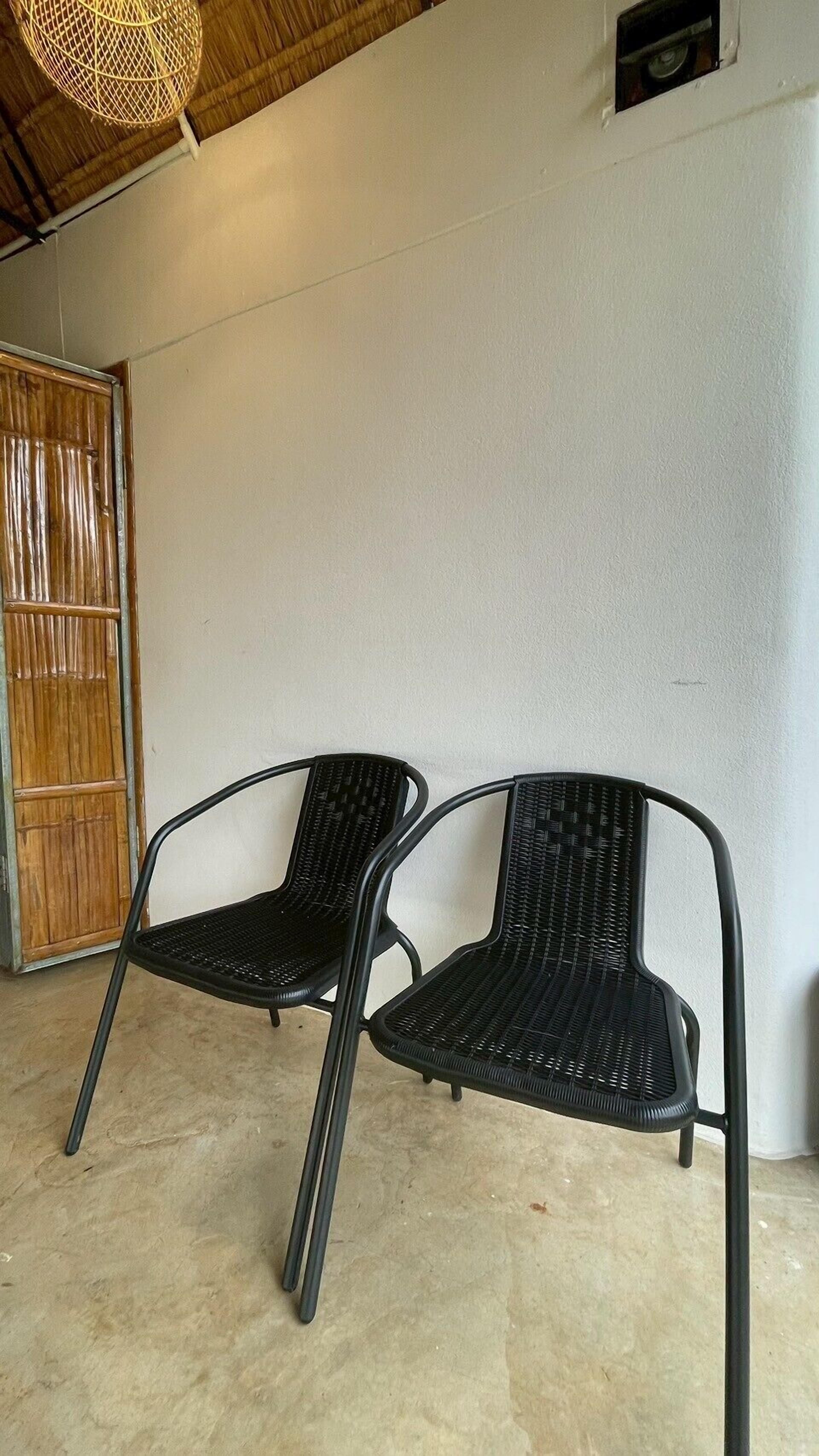 Due sedie nere in vimini con struttura metallica, posizionate di fronte a una parete bianca in una stanza con pavimento in cemento e porta in legno.