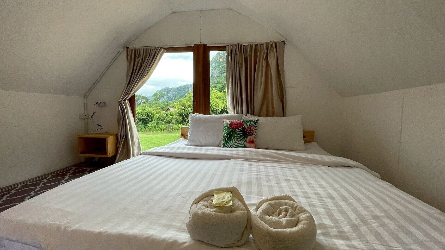 A bedroom with a large bed, a nightstand, a window with curtains, and a view of greenery outside.