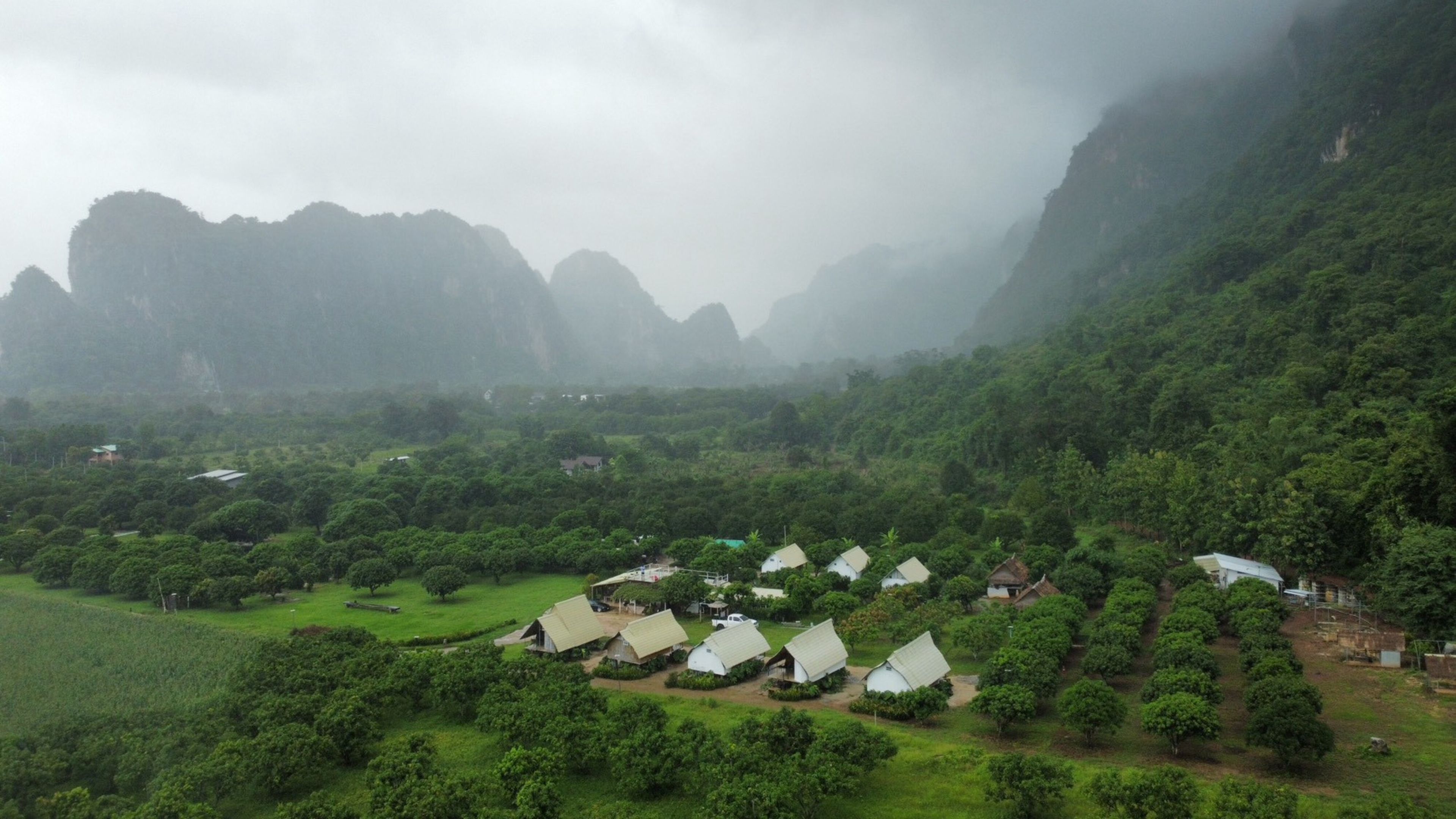 A rural landscape with several white-roofed buildings surrounded by lush greenery and mountains in the background.