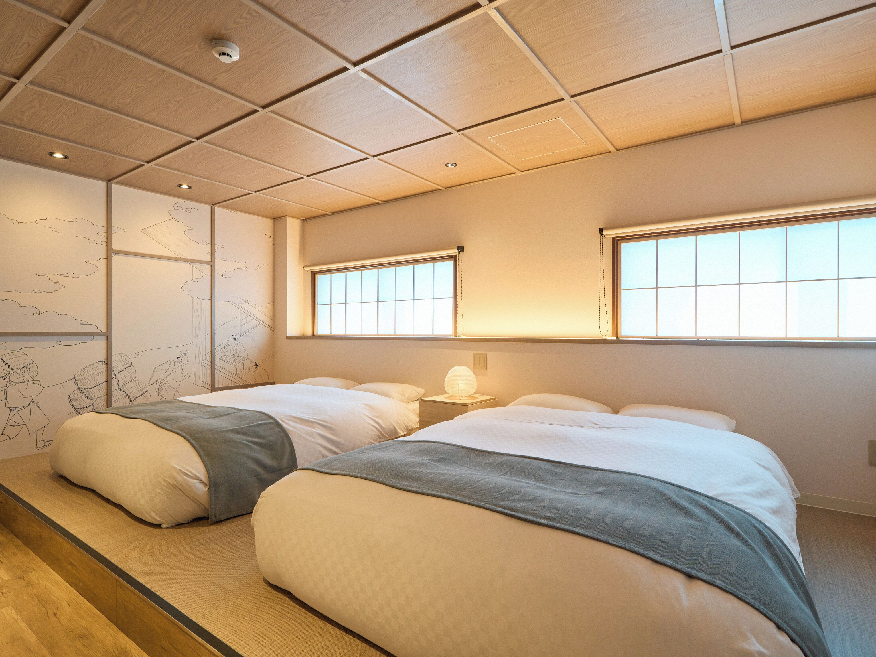 A hotel room with two beds, a wooden floor, a wall with a mural, and a window with blinds.