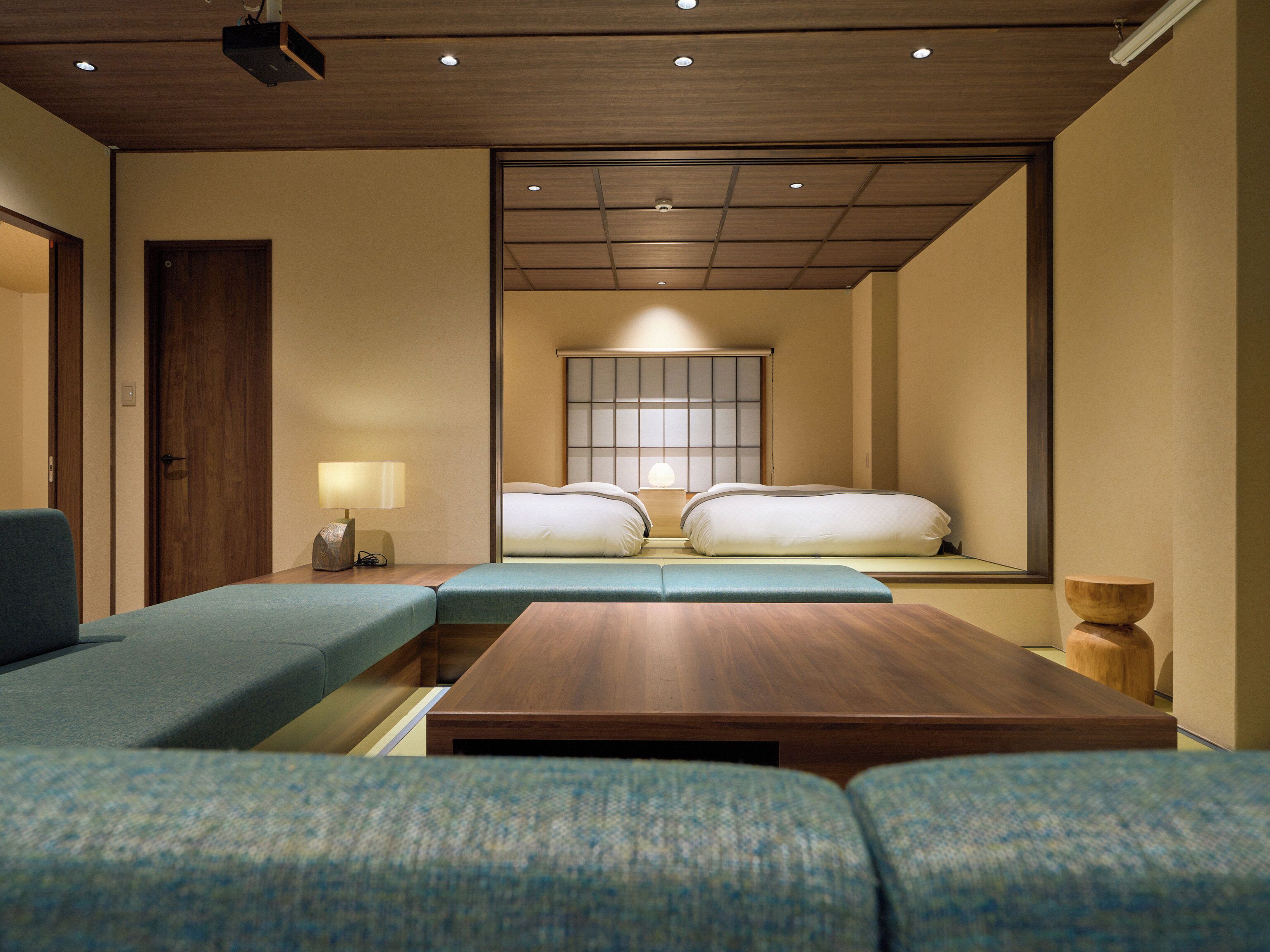 Una habitación de hotel moderna con un sofá, una mesa de centro de madera, una mesita de noche con lámpara y una tradicional pantalla shoji japonesa detrás de la cama.
