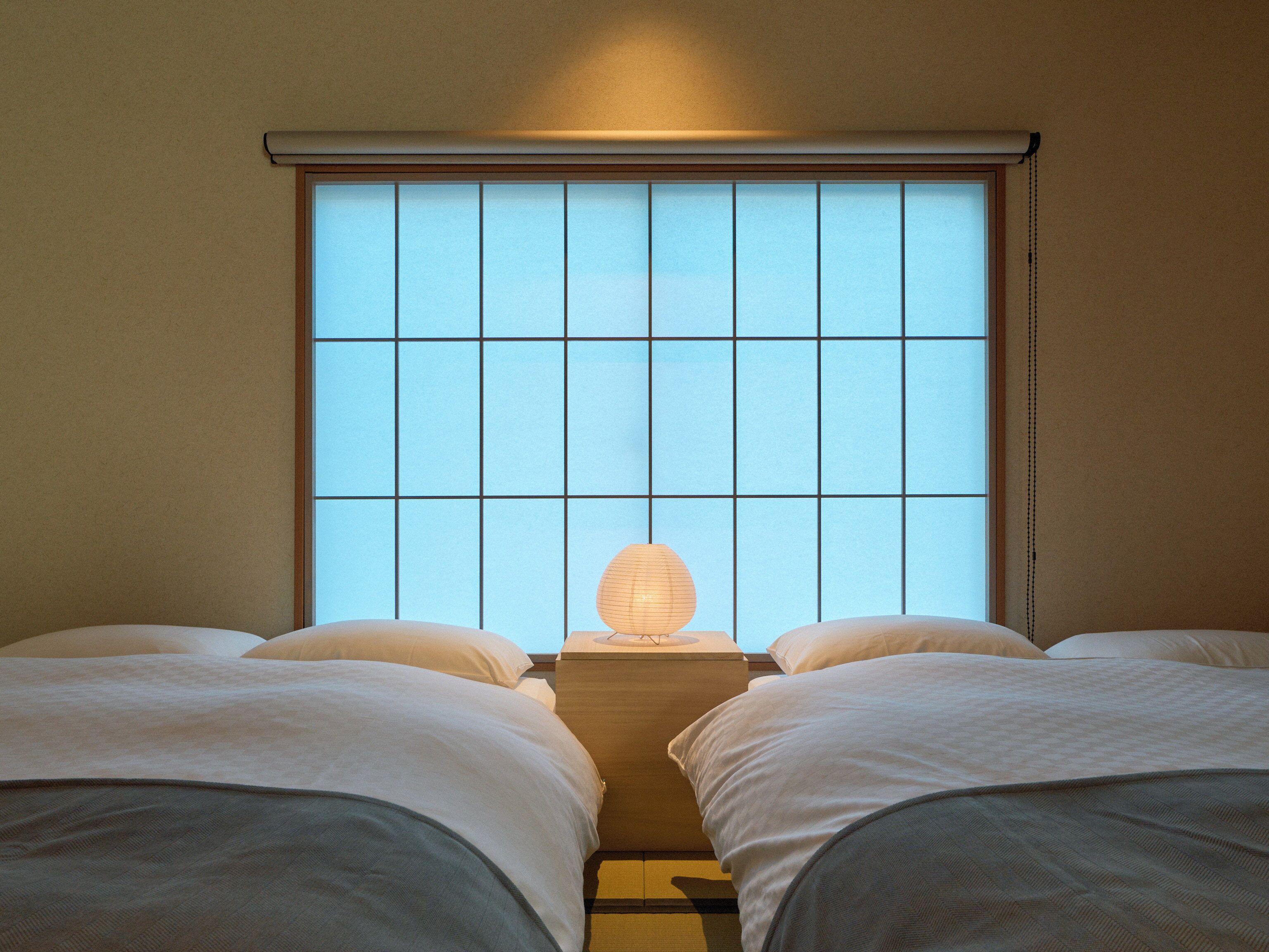 Een kamer met twee bedden, een raam met een roedeverdeling en een nachtkastje met een lamp.