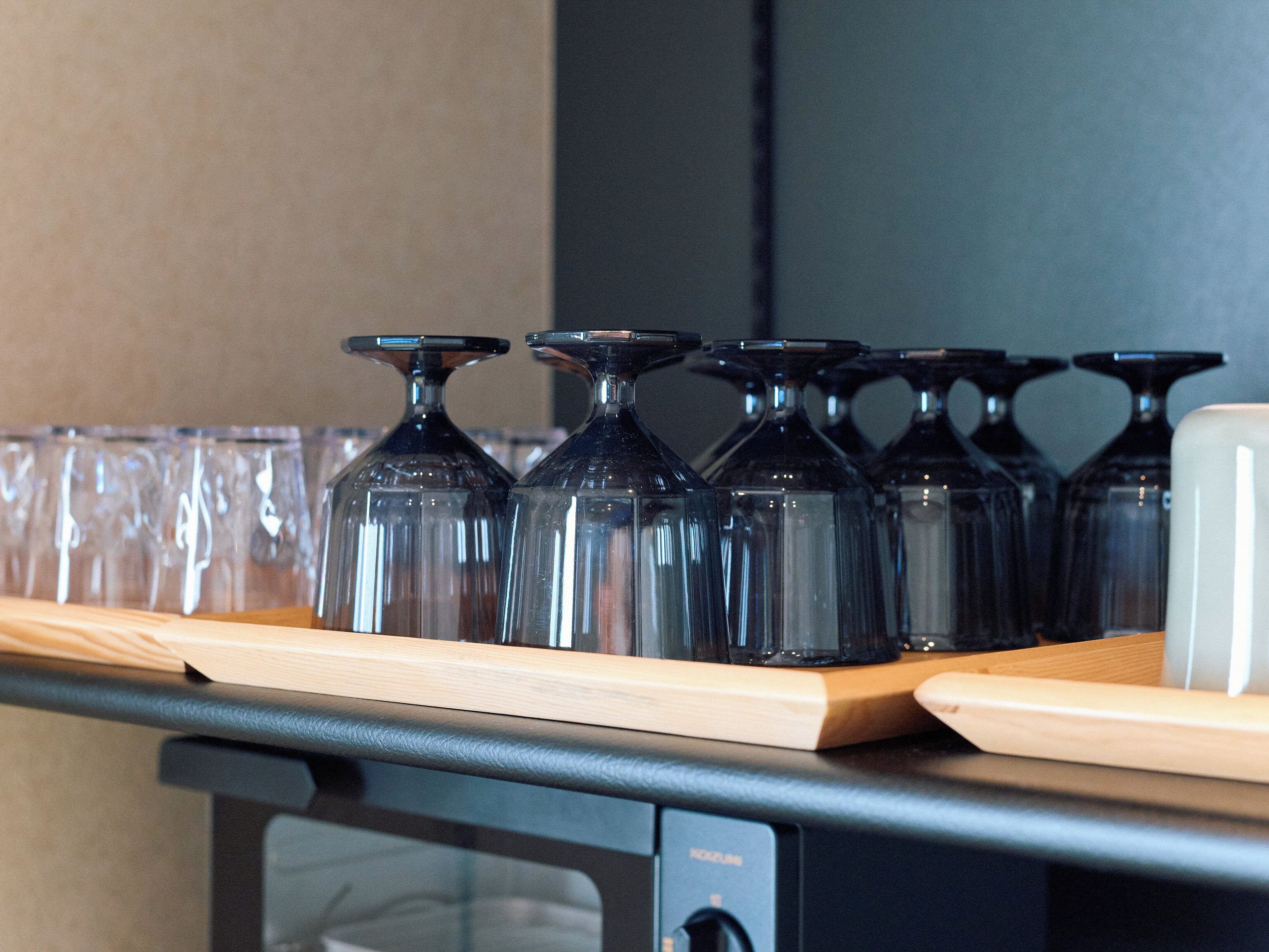 A row of clear wine glasses on a wooden shelf above a black appliance, likely a refrigerator.
