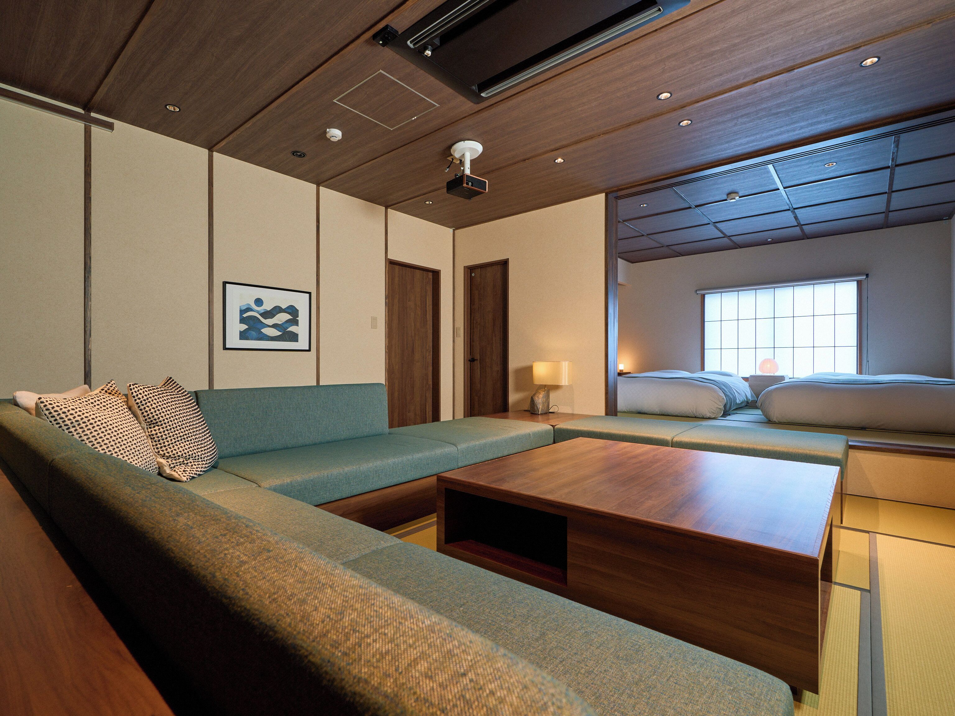 A modern hotel room with a sofa, a wooden coffee table, a bed with a view, and a ceiling with recessed lighting.