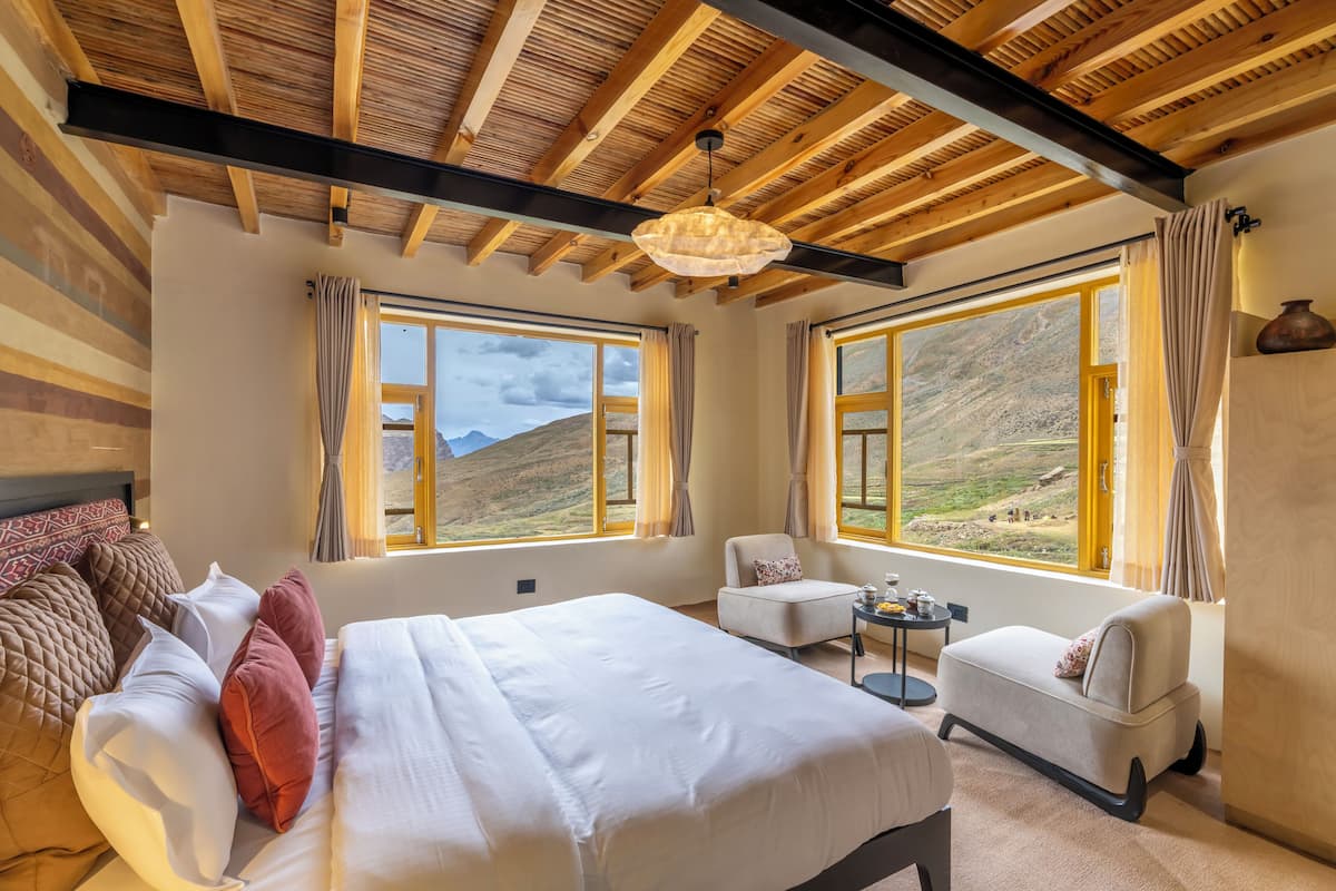 A room with a large bed, two armchairs, and a small table. There are large windows with curtains, a wooden ceiling, and a hanging light fixture.