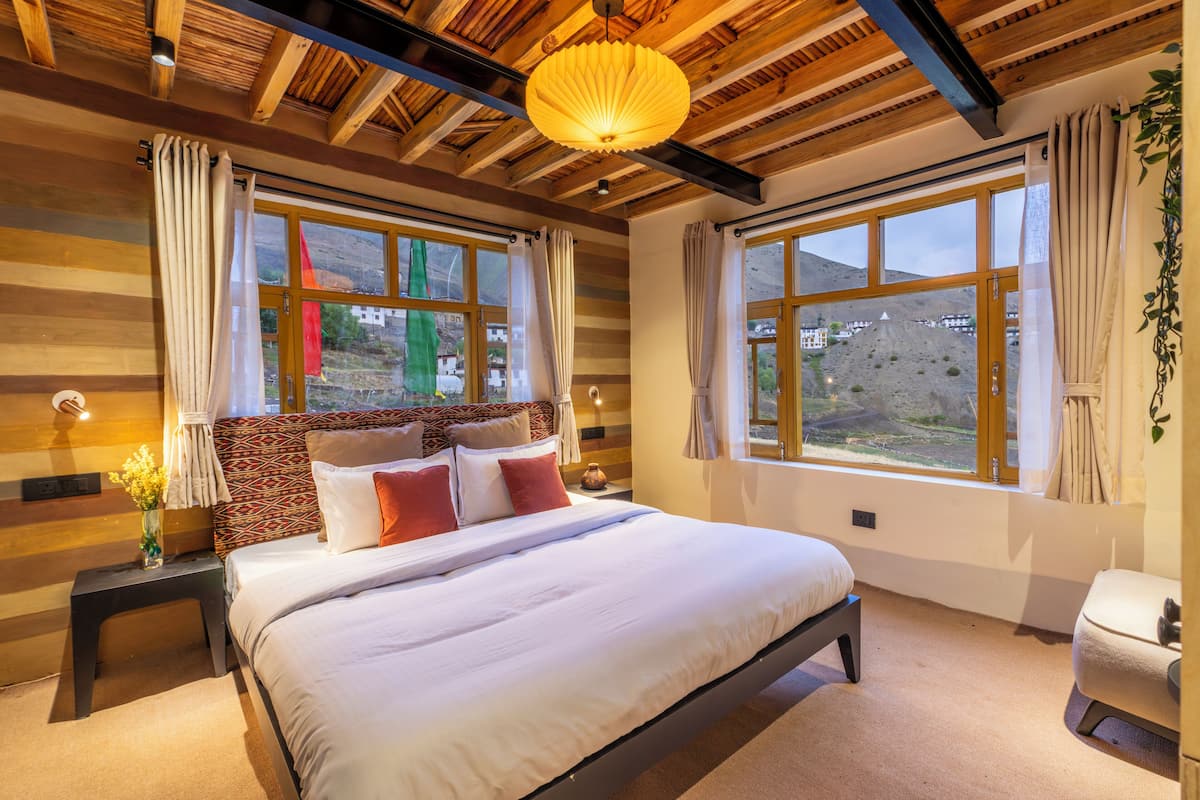 A bedroom with a large bed, wooden ceiling beams, and a view of a mountainous landscape through large windows.