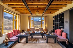 A dining area with a view of mountains, wooden ceiling beams, and a bookshelf. - House on the Clouds (Spiti)