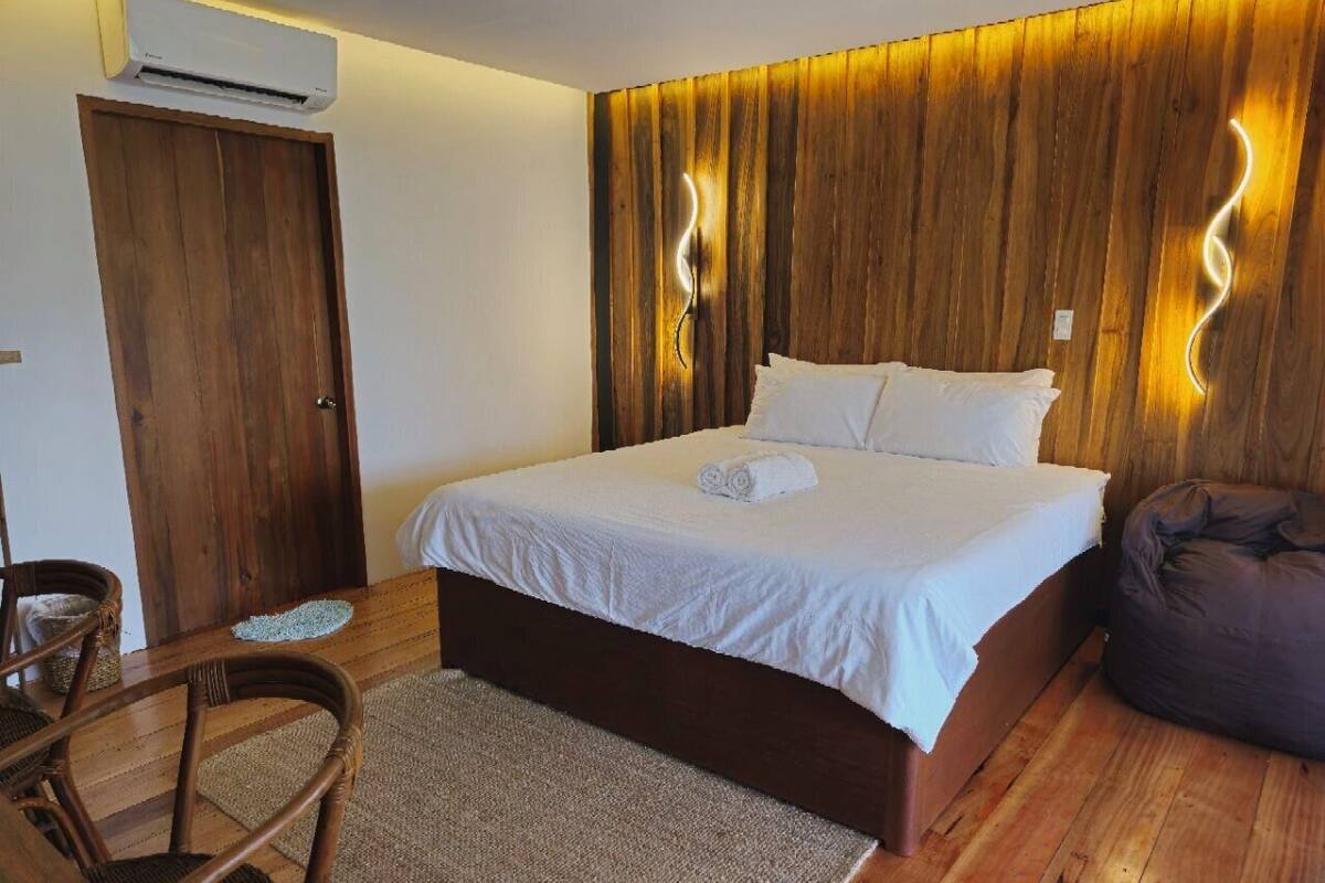 Un dormitorio con cabecera de madera, cama con ropa de cama blanca, una silla y un sillón de felpa.