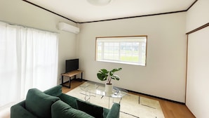 A living room with a green sofa, a glass coffee table, a potted plant, a TV on a stand, and a window with a view of greenery.