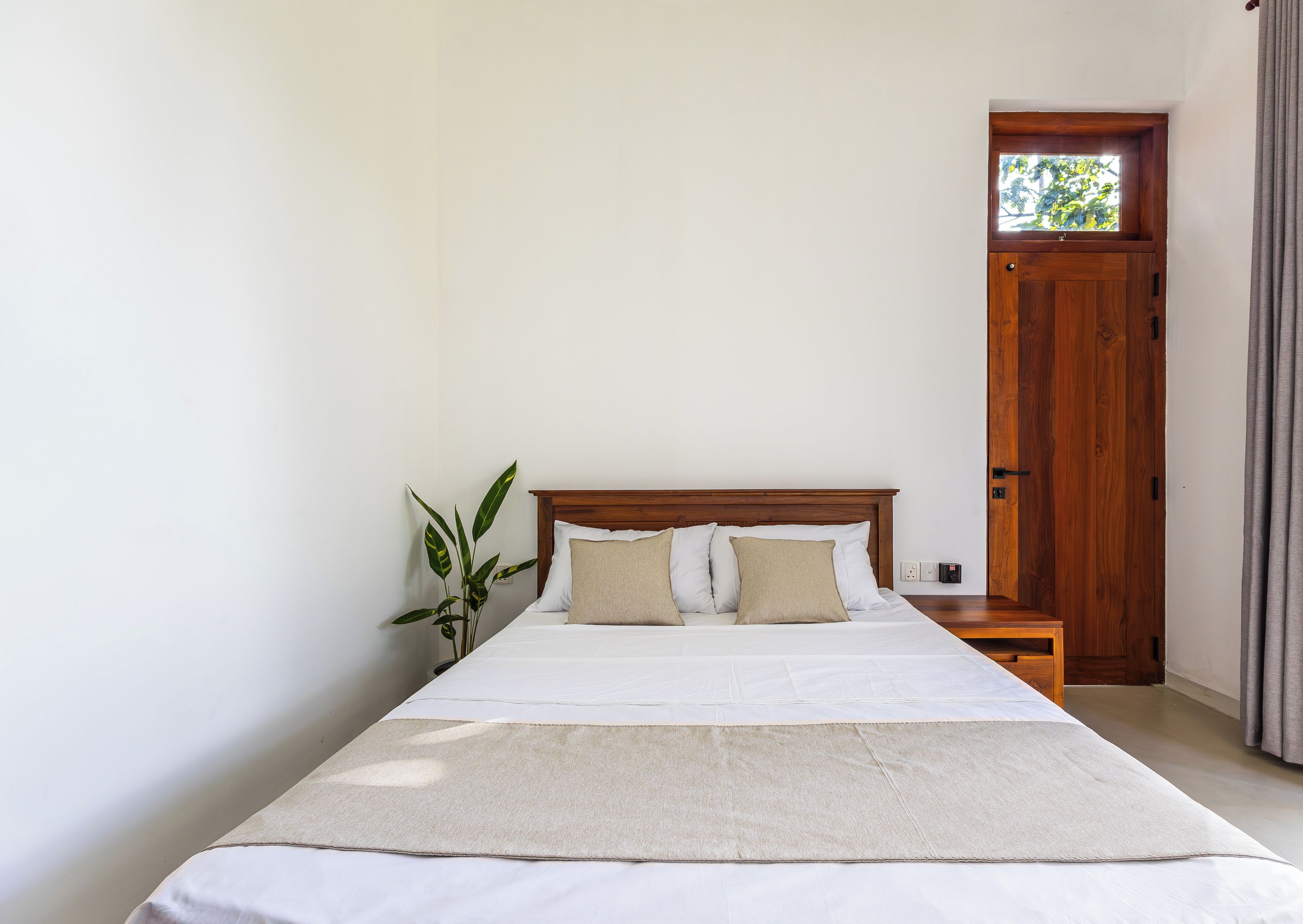 Quarto com cama, mesa de cabeceira, porta de madeira e vista para o exterior.