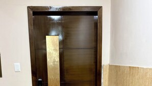 Una puerta marrón con picaporte dorado y mirilla, que da a un pasillo con suelo de baldosas y felpudo.