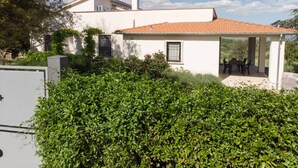 Ein weißes Haus mit Ziegeldach, einer überdachten Terrasse mit Gartenmöbeln und üppiger Bepflanzung im Vordergrund.