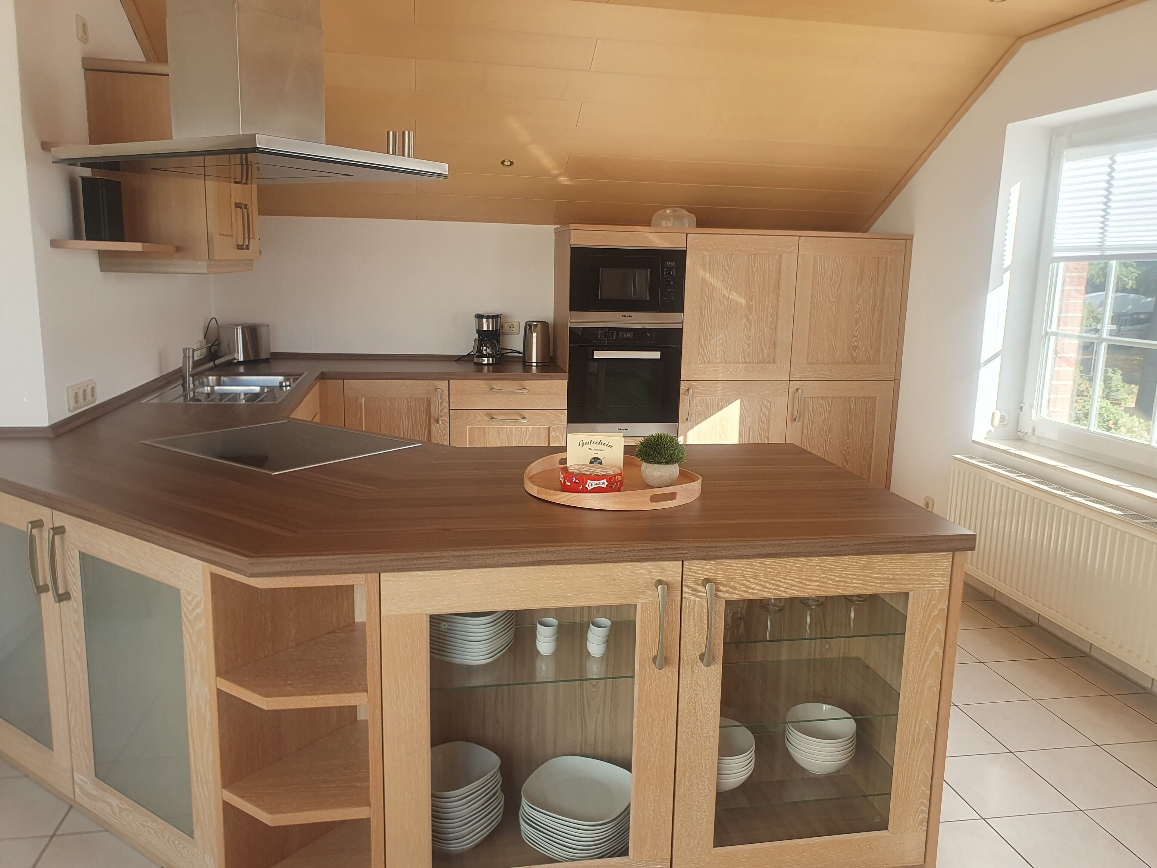 Eine moderne Küche mit Holzschränken, einem eingebauten Ofen und einer großen Insel mit Glastür, hinter der Teller ausgestellt sind.