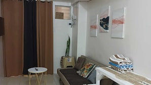 A small, well-lit living room with a sofa, a small round table, and a white bench. There are curtains, a plant, and a wall-mounted TV. - Cozy City Stay in Baguio (Baguio)