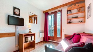 A cozy room with a television, a mirror, a shelf with decorative items, a bed with pink bedding, and a window with a view of trees.