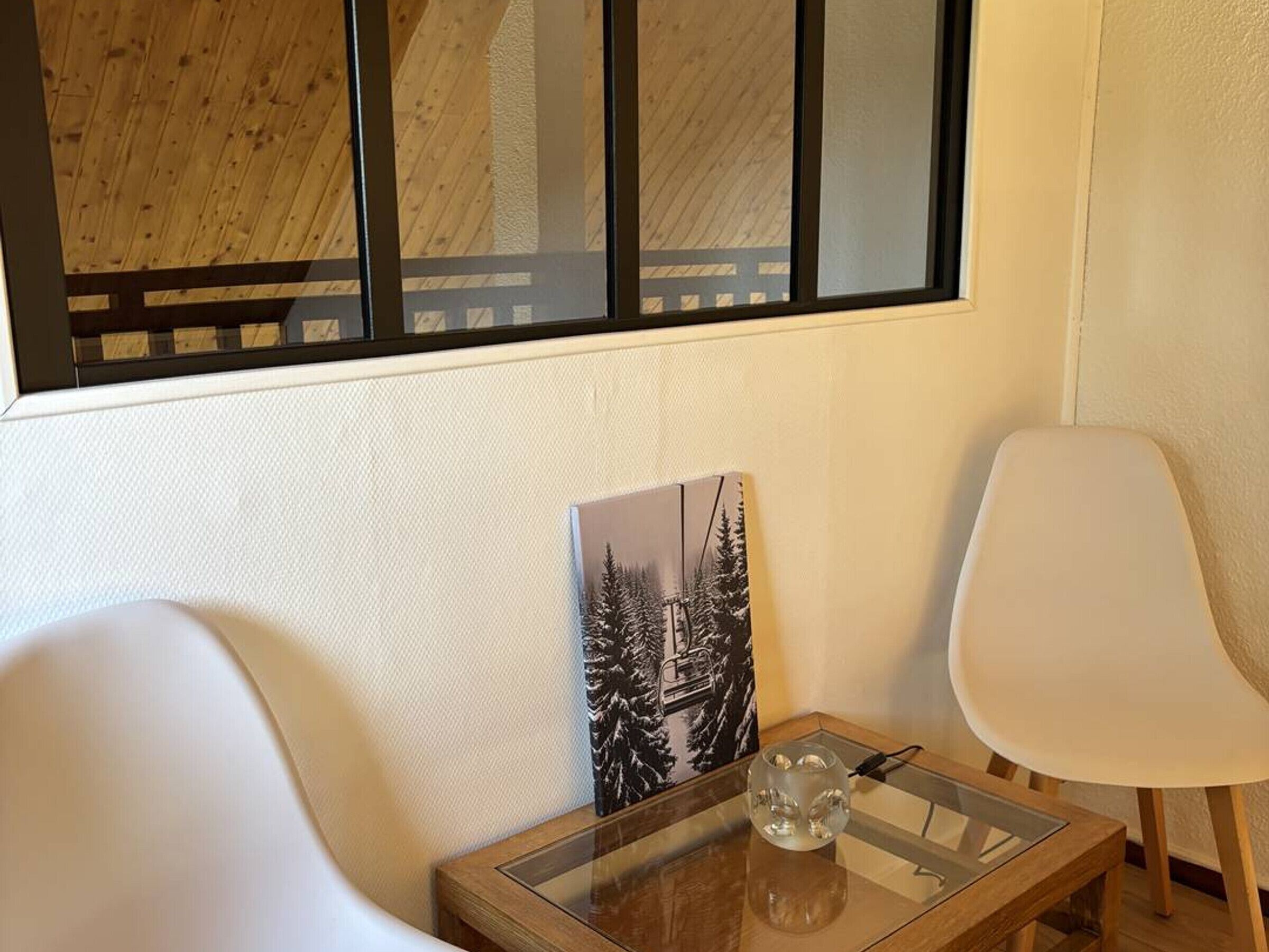 A room with a wooden ceiling, a window with a black frame, a white wall, a glass table with a clear glass object, and two white chairs.