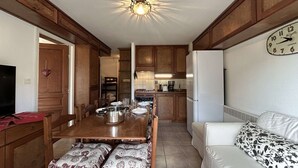A compact kitchen with wooden cabinets, a dining table set for four, a white sofa, and a wall clock.