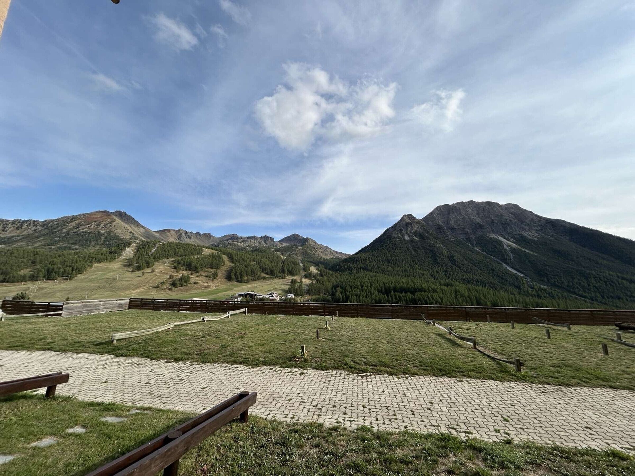 Un sentier pavé bordé de bancs, une zone herbeuse et, au loin, une chaîne de montagnes.