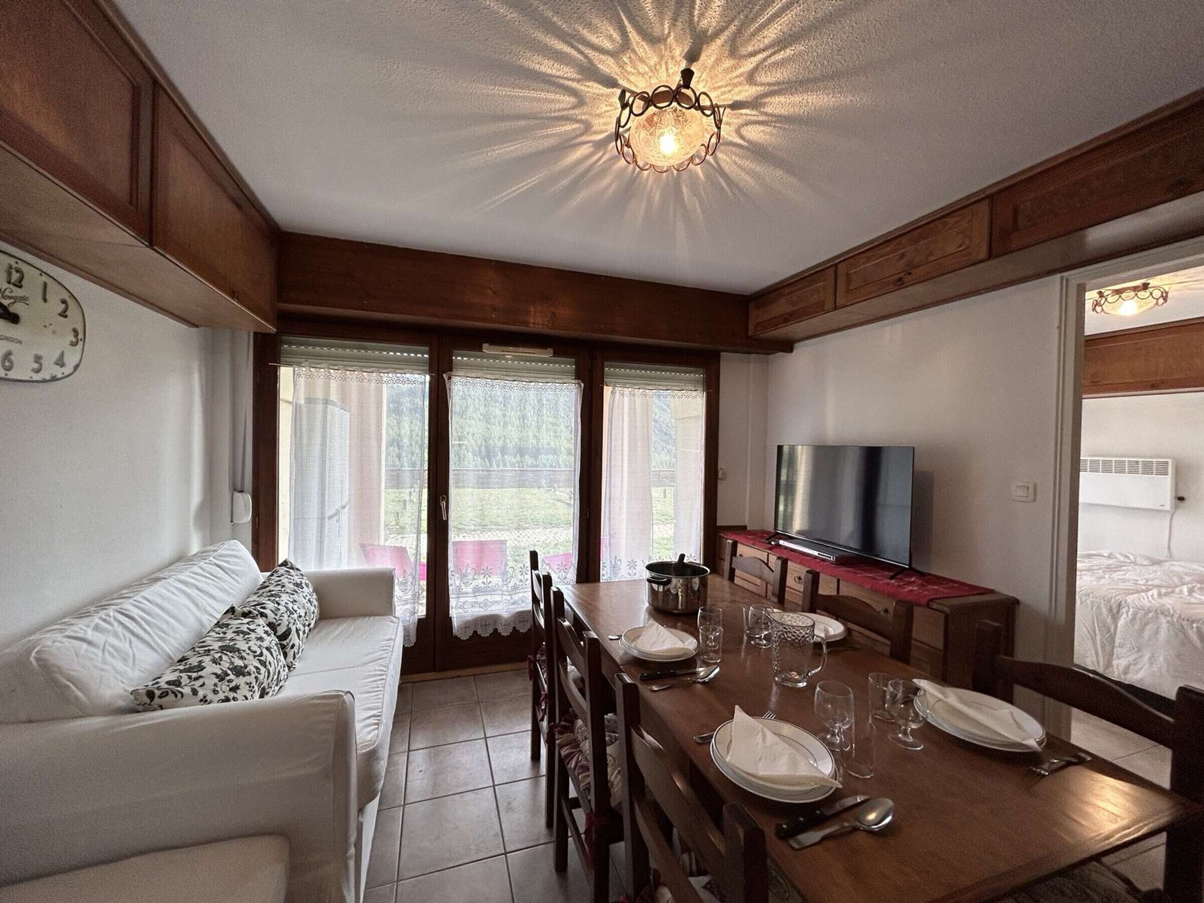 Une pièce confortable avec un canapé, une table à manger dressée pour quatre personnes, un téléviseur et une grande fenêtre avec des rideaux.